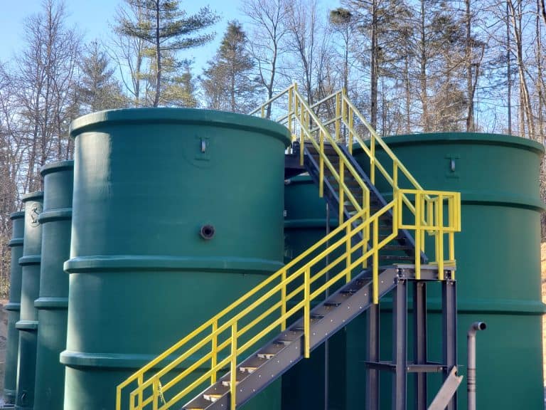 Large green industrial storage tanks with yellow safety stairs in an outdoor facility, surrounded by trees, showcasing A3 USA's water and wastewater treatment solutions.